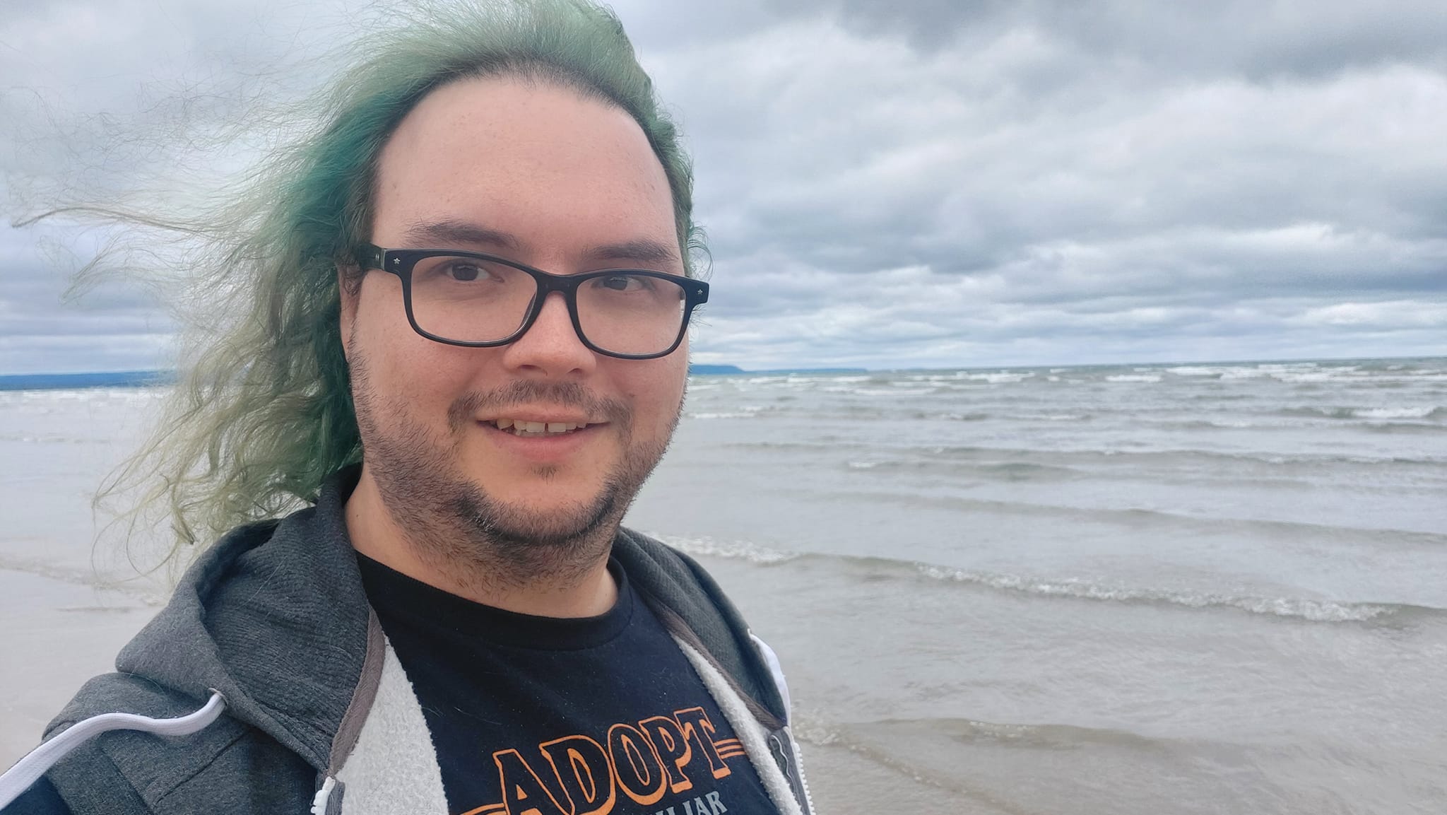 A man with long green hair stands on a beach, the water is choppy and the sky cloudy